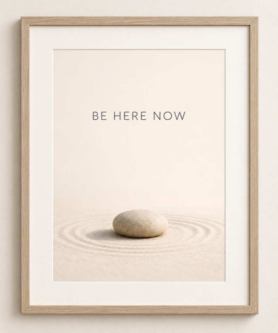 A framed print reading Be Here Now, with a smooth stone resting on raked sand