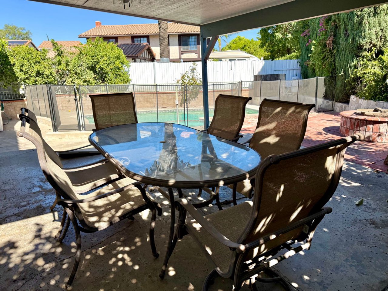 A shaded outdoor dining table with six chairs, looking out at the fenced pool