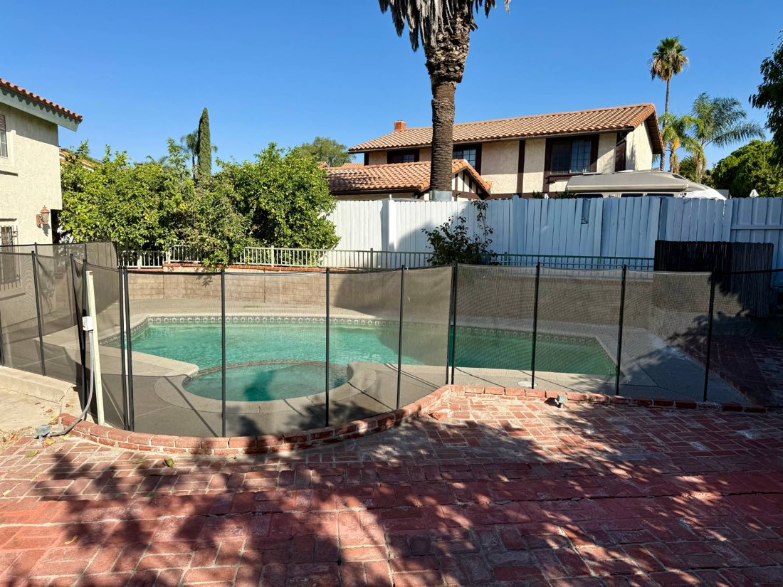 The backyard, with a brick patio and a fenced pool under palm trees